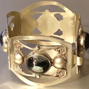 Puede incluir: Una pulsera de pu&ntilde;o plateada con un dise&ntilde;o ornamentado. La pulsera presenta una piedra oscura ovalada central, rodeada de piedras redondas m&aacute;s peque&ntilde;as y volutas decorativas. La parte superior tiene formas recortadas.