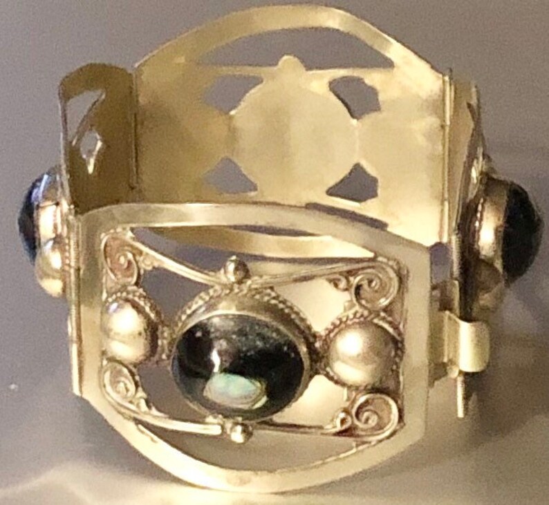 Puede incluir: Una pulsera de pu&ntilde;o plateada con un dise&ntilde;o ornamentado. La pulsera presenta una piedra oscura ovalada central, rodeada de piedras redondas m&aacute;s peque&ntilde;as y volutas decorativas. La parte superior tiene formas recortadas.