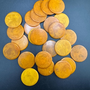 May include: A collection of round, amber-colored game pieces with a marbled pattern. The pieces vary in shades of yellow and brown, scattered on a dark surface. The game pieces are likely for a board game or other tabletop activity.