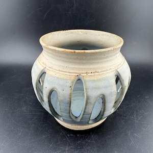 May include: A handmade ceramic vase in light blue and cream. The vase has oval cutouts around the body. The interior is blue, and the rim is a lighter cream colour. The vase is a decorative item.