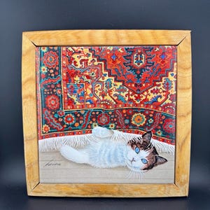 May include: A square wooden frame containing a print of a white cat with blue eyes lying on a light-coloured floor in front of a vibrant, multi-coloured patterned rug with red, blue, and orange details. The artist's signature is visible in the corner.