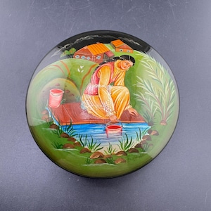 May include: Round, hand-painted decorative box with a detailed scene. The image depicts a person in yellow clothing near a body of water, with a red bucket. The background features green hills, houses, and foliage. The box has a glossy finish.