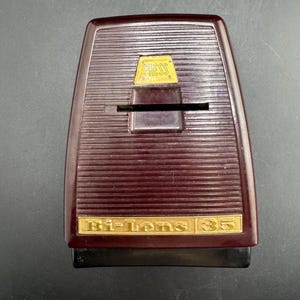 May include: A dark maroon plastic slide viewer with a horizontal ribbed texture. It features a gold-colored rectangular plate at the top with the word PRESS embossed in black, and a gold-colored plate at the bottom labeled Bi-Lens 35.