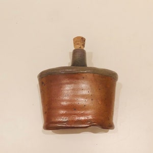Może przedstawiać: Brązowa ceramiczna butelka z korkowym korkiem. Butelka ma lekko nierówny, ręcznie robiony wygląd.