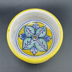 May include: A small, round ceramic bowl with a bright yellow rim and a floral design. The interior features a yellow circle with a blue and green floral pattern. The bowl's interior is white, and the design is centered.