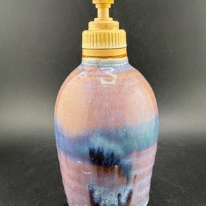 May include: A ceramic soap dispenser with a tan pump and a rounded body. The body features a blend of pink, blue, and brown hues, creating a marbled effect. The dispenser is designed for liquid soap.