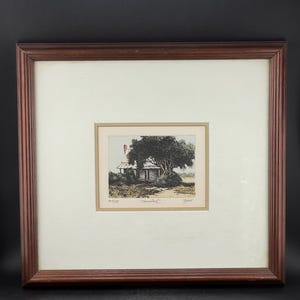 May include: A framed print titled Homestead featuring a small house partially obscured by a large tree. The artwork is matted in off-white and enclosed in a dark brown wooden frame with vertical ribbed detailing along the outer edge.