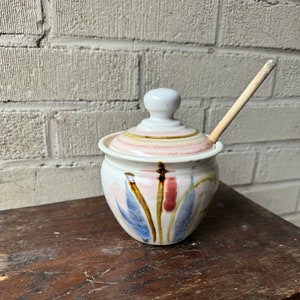 May include: A white ceramic honey pot with a lid and a wooden honey dipper. The pot has a floral design in shades of blue, pink, and brown.