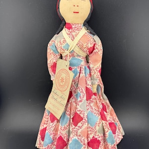 Może przedstawiać: Ręcznie robiona lalka z materiałową sukienką z wzorem czerwonych, niebieskich i różowych rombów. Lalka ma czarne warkocze, czerwoną opaskę i metkę. To przedmiot vintage.