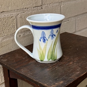 May include: White ceramic mug with a blue band and a hand-painted design of blue irises with green leaves.