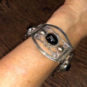 Puede incluir: Pulsera plateada con un dise&ntilde;o ornamentado. La pulsera presenta una gran gema negra ovalada en el centro, rodeada de peque&ntilde;os detalles plateados y piedras negras adicionales. La pulsera est&aacute; en una mu&ntilde;eca.