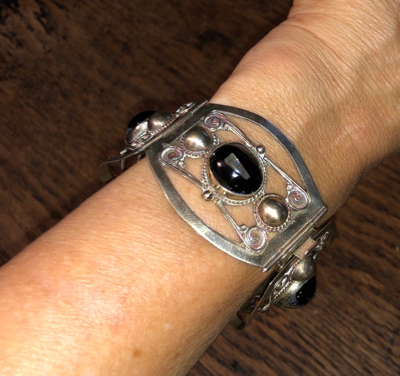 Puede incluir: Pulsera plateada con un dise&ntilde;o ornamentado. La pulsera presenta una gran gema negra ovalada en el centro, rodeada de peque&ntilde;os detalles plateados y piedras negras adicionales. La pulsera est&aacute; en una mu&ntilde;eca.