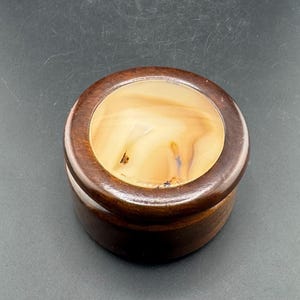 Puede incluir: Una caja redonda de madera con una tapa de piedra de color marrón claro y crema. La caja está hecha de madera marrón oscuro y tiene un acabado liso y pulido. La tapa de piedra tiene un patrón único y arremolinado.