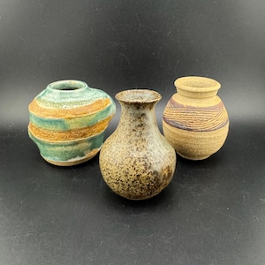 May include: Three small, handcrafted ceramic vases. The vases are in shades of green, brown, and tan. One vase has horizontal stripes, another has a speckled pattern, and the third has a textured design.