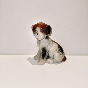 Könnte beinhalten: Eine Keramikfigur eines braunen und weißen Hundes, der mit gesenktem Kopf sitzt. Der Hund hat braune Augen und eine schwarze Nase.