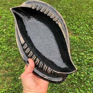 May include: Black ceramic dish with a gold rim and a leaf design. The dish is oval shaped and has a glossy finish.