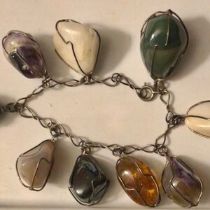 Pode incluir: Um bracelete de corrente de prata com oito pedras de cores diferentes, cada uma envolta em uma gaiola de arame. As pedras incluem ametista, &aacute;gata branca, &aacute;gata verde, hematita e &acirc;mbar.