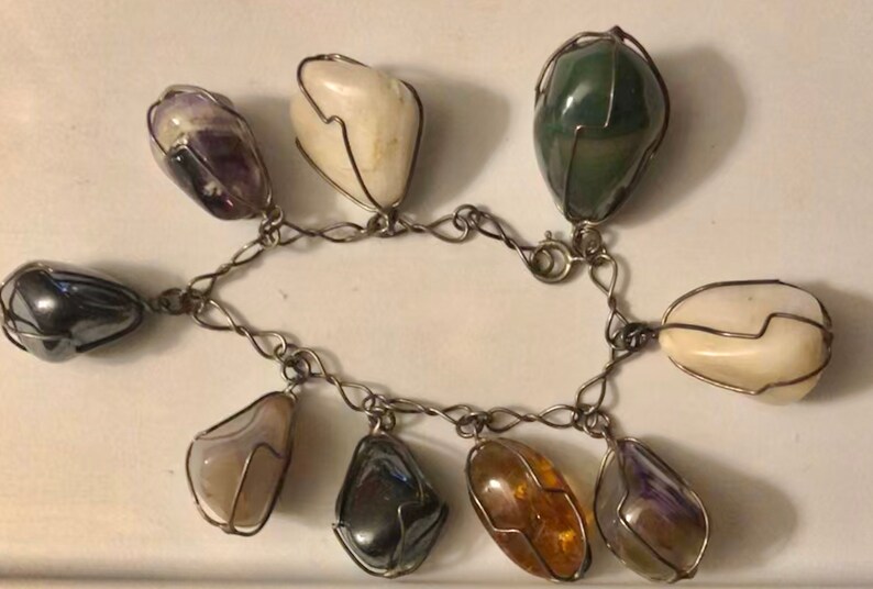 Pode incluir: Um bracelete de corrente de prata com oito pedras de cores diferentes, cada uma envolta em uma gaiola de arame. As pedras incluem ametista, &aacute;gata branca, &aacute;gata verde, hematita e &acirc;mbar.
