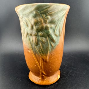 Puede incluir: Un jarrón de cerámica con la parte superior acampanada y una base redondeada. El jarrón presenta un diseño texturizado con tonos marrones y verdes. El diseño parece ser de temática floral o botánica. El jarrón es un objeto decorativo.