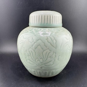 May include: A celadon-colored ceramic jar with a matching lid. The jar features a floral pattern and a ribbed lid. The jar is a light green color and has a rounded shape. The jar is likely used for storage or decoration.