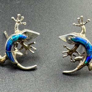 Puede incluir: Un par de pendientes en forma de lagartija. Cada lagartija presenta un cuerpo curvado con incrustaciones de material azul iridiscente que muestra reflejos verdes y turquesas. El metal tiene un acabado pulido con patas detalladas.