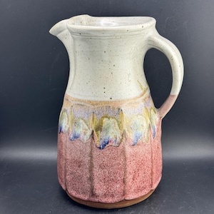 May include: A ceramic pitcher featuring a speckled off-white upper section and a textured, fluted lower body in shades of dusty rose. The transition between the two sections displays a decorative drip glaze in muted tones of blue, green, and gold.