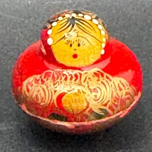 May include: A small, round, red decorative object featuring a painted yellow face with dark hair, white dots, and a red mouth. The lower portion displays intricate, swirling gold-colored line patterns across the vibrant red surface.