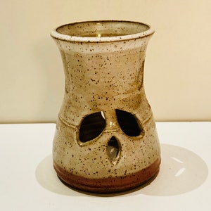 May include: A white ceramic oil burner with a speckled glaze and a red rim. The burner has a unique design with cut-out holes in the shape of a face.