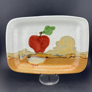 May include: Rectangular ceramic serving dish with a hand-painted apple design. The dish features a red apple with a green leaf, set against a white and tan background. The rim is a speckled white, and the base is a warm brown.