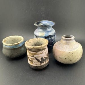May include: Four handcrafted ceramic vases in various shapes and sizes. The vases feature a range of colors, including blue, gray, and beige, with unique textured finishes. The collection includes a small, round vase and a taller, cylindrical one.