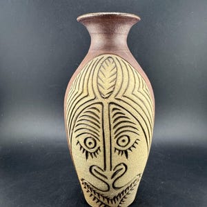 May include: A brown ceramic vase with a carved face design. The vase has a narrow neck and a wider body, with a detailed face carved into the front. The face features large eyes, a nose, and a smiling mouth. The vase is approximately 10 inches tall.