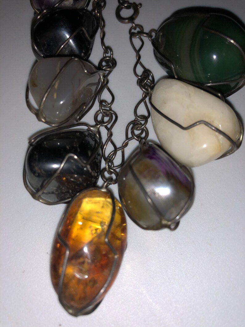 Pode incluir: Um bracelete de corrente de prata com sete pedras de diferentes cores, cada uma envolta em arame. As pedras incluem &acirc;mbar, ametista, &aacute;gata, verde, preto e branco.