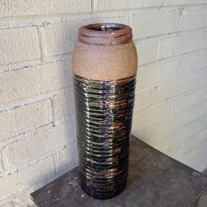 Puede incluir: Un jarrón cilíndrico de cerámica, con una sección superior texturizada y marrón y una sección inferior negra y acanalada. El jarrón tiene un interior oscuro y se apoya sobre una superficie oscura. Cerámica hecha a mano para la decoración del hogar.