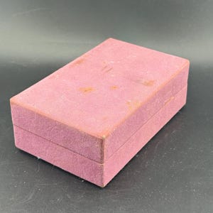 May include: A rectangular, dusty rose-colored box with a velvet-like texture. The box is closed, with a lid that fits snugly over the base. The box appears to be vintage, with some discoloration on the top surface.