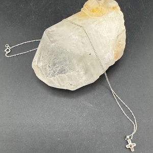 Op de afbeelding: Een zilveren ketting met een kruisvormige hanger met een kleine rode edelsteen. De ketting wordt getoond op een groot, helder bergkristal tegen een donkere achtergrond.