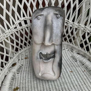 May include: A white and gray ceramic sculpture of a face with exaggerated features. The face has a large nose, wide eyes, and a gaping mouth.