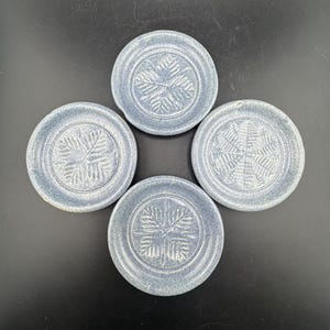 May include: Four round, blue-grey ceramic coasters. Each coaster features a raised, symmetrical leaf design in the center. The coasters are arranged in a square formation, showcasing their decorative and functional nature.