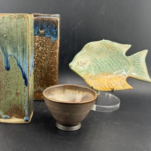 May include: A collection of handmade ceramic pieces. Includes two rectangular vases with green, blue, and brown glaze, a small brown bowl, and a fish-shaped plate with green and yellow glaze.