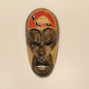 May include: A wooden mask with a carved face and intricate designs. The mask is painted in shades of brown, black, and gold. A red and yellow turtle is painted on the forehead.