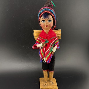 May include: A vintage doll dressed in traditional attire, featuring a red poncho with colorful trim, black shorts, and a patterned hat. The doll carries a small bundle on its back and stands on a wooden base.