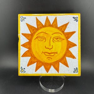May include: Square ceramic tile with a smiling sun design in yellow and orange. The sun has a face with eyes, a nose, and a mouth. The tile has a white background, a yellow border, and small blue accents. The tile measures approximately 15 cm x 15 cm.