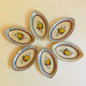 Puede incluir: Seis pequeñas bandejas ovaladas de cerámica blanca con ribetes azules y dorados. Cada bandeja presenta un diseño floral con flores amarillas, rojas y rosas.