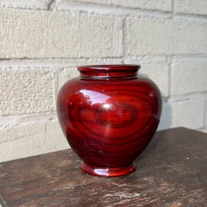 May include: A small, round, red wooden vase with a glossy finish. The vase is made of wood with a natural grain pattern.