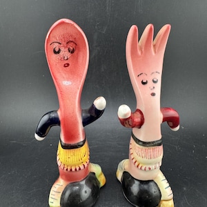 Op de afbeelding: Een paar vintage keramische zout- en peperstrooiers in de vorm van antropomorfe lepel- en vorkfiguren. De lepel en vork hebben rode gezichten en lichamen, met zwarte en witte accenten op hun armen en benen. De lepel en vork staan op zwarte voetstukken.