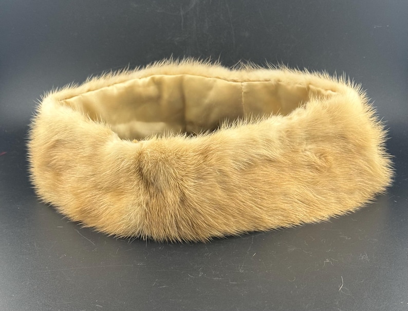 May include: A circular fur collar in light brown, featuring a soft, pale lining. The fur has a dense, plush texture. The collar is designed to be worn around the neck. The item is likely a fashion accessory.