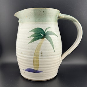 Puede incluir: Una jarra de cerámica blanca con un diseño de palmera verde. La jarra tiene un esmalte verde claro en la parte superior y una base azul.
