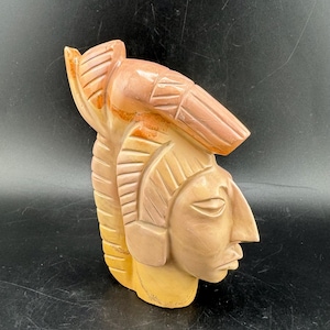 Könnte beinhalten: Eine geschnitzte Skulptur eines indianischen Kopfes mit Kopfschmuck und detaillierten Gesichtszügen. Die Skulptur ist hauptsächlich beige mit rotbraunen Akzenten und weist aufwendige Schnitzereien und eine glatte Oberfläche auf.