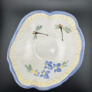 May include: Handmade ceramic bowl with a light blue rim and a white interior. The bowl is decorated with blue flowers, green leaves, and two dragonflies. The bowl has a unique, irregular shape.
