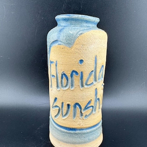 May include: A ceramic vase with a light blue and beige glaze. The vase has the words "Florida Sunshine" written on it in blue.