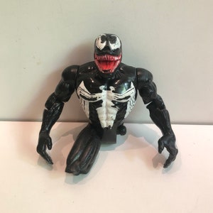 May include: Black and white Venom action figure with a red mouth. The figure is in a crouching pose with its arms outstretched.